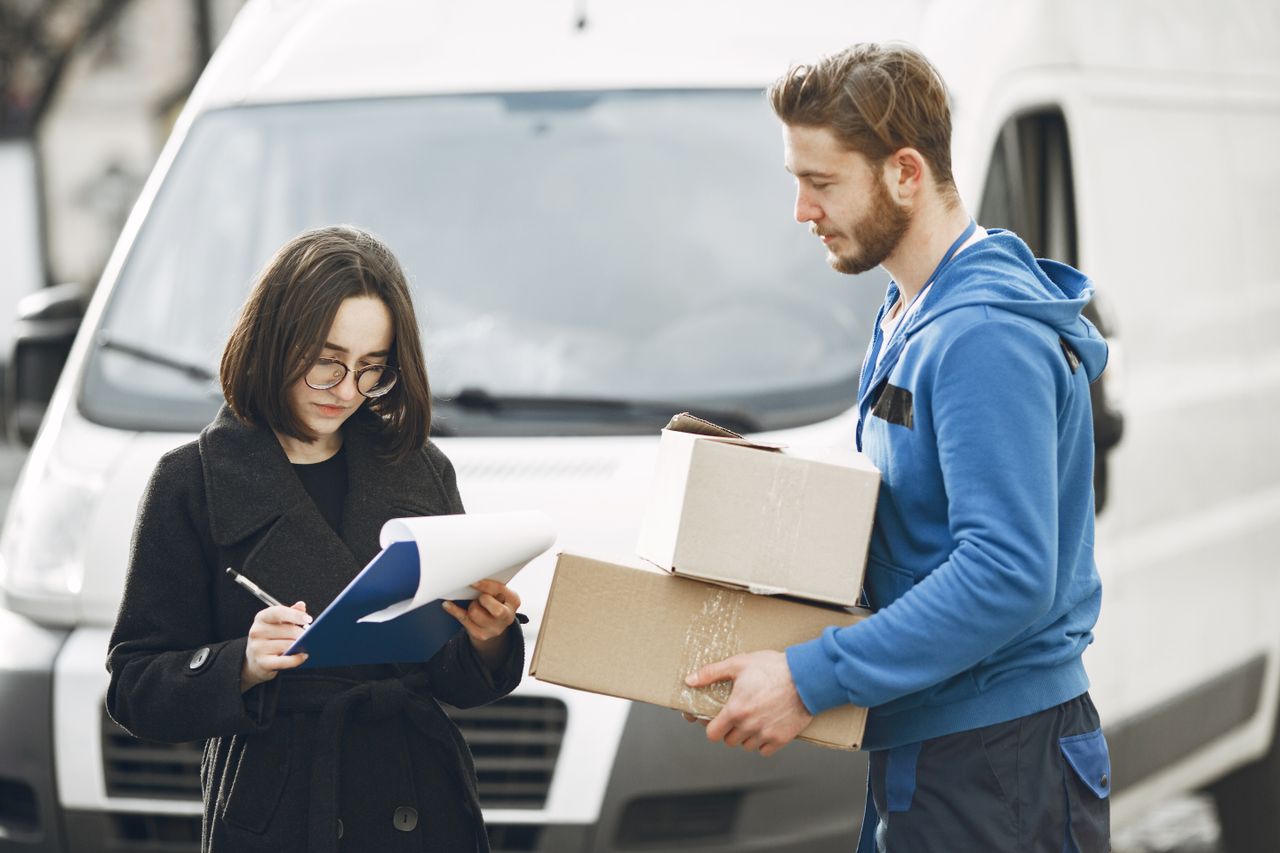 hero-img-01 A courier in blue hands packages to a woman signing documents in front of a delivery van.