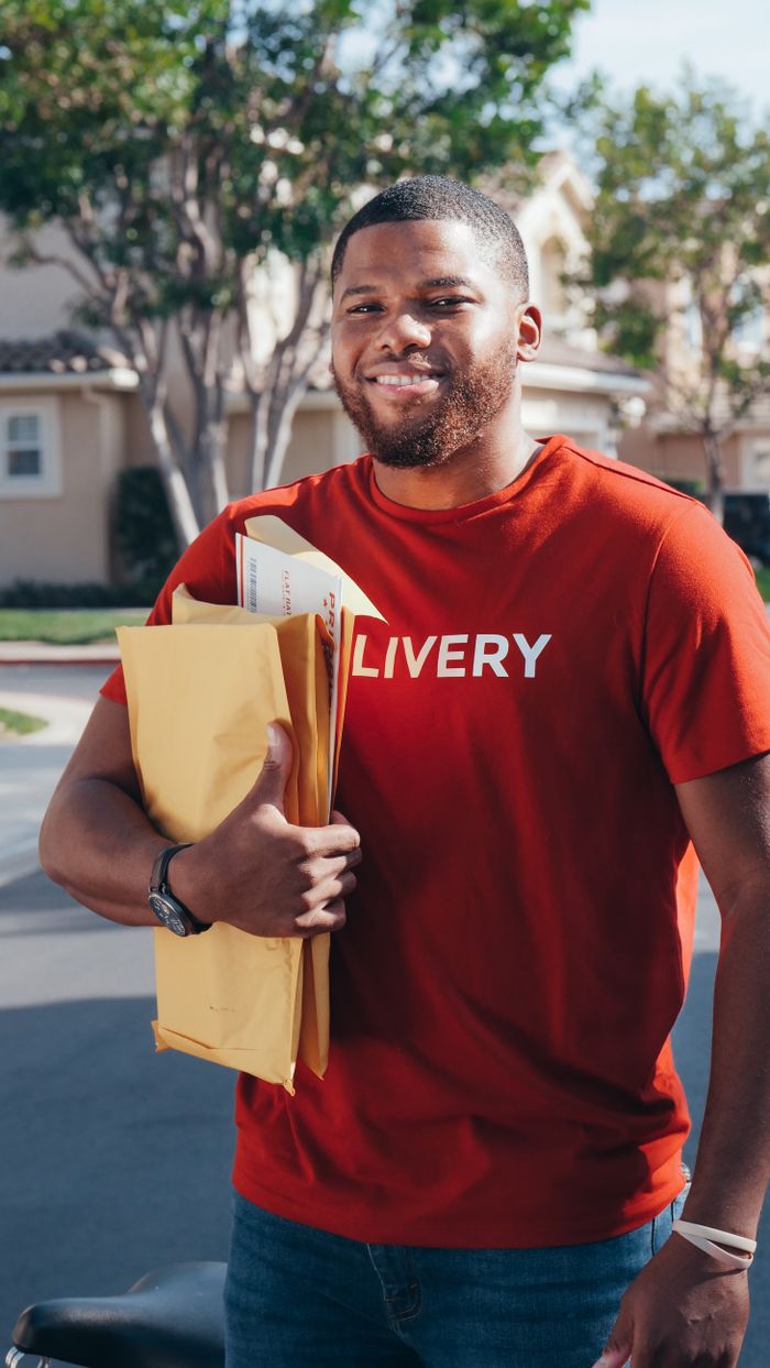 gallery-3 Cheerful delivery man holding packages in a sunny neighborhood setting.