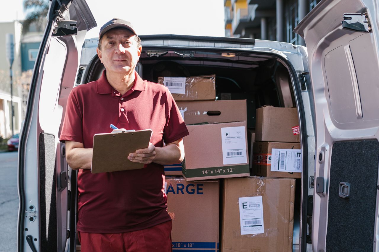 about-01 Elderly delivery person holds clipboard in front of open van filled with packages on a sunny day.