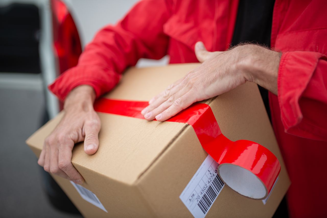 hero-img-02 A courier in red uniform sealing a cardboard package with red tape for delivery.