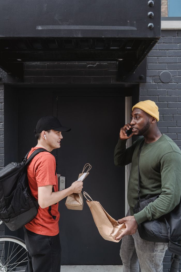 gallery-1 Two men exchange delivery bags at a building entrance, one on the phone.